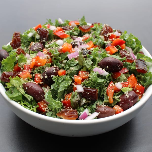 A vibrant Garden Kitchen kale salad with olives, red peppers, red onions, and dates in a white bowl.